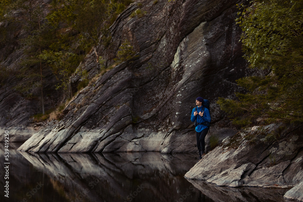 A caucasian man standing on a rock with hiking gear by a lake in a forest.