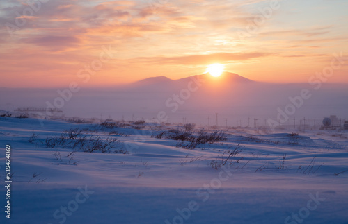 Winter arctic landscape. Industrial structures, electrical pylons and a satellite dish in the winter snow-covered tundra in the Arctic. Sunset over the tundra and mountains. Chukotka, Siberia, Russia.