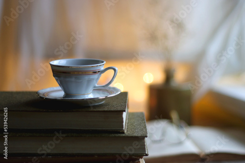 Plate of chocolate chip cookies, stack of vintage books, reading glasses, cup of tea or coffee, lit candle and fairy lights. Hygge at home. Selective focus.