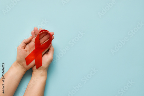Healthcare and medicine concept - female hands holding red AIDS awareness ribbon on blue background. The symbol of The World AIDS Day or cancer or HIV Awareness Month. Top view with copy space 