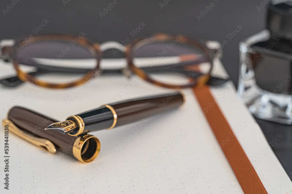 Close up photo of pen and blurred glasses, ink bottle and planner on a gray background. Selective focus Close up photo of pen and blurred glasses, ink bottle and planner on a gray background. Selective focus