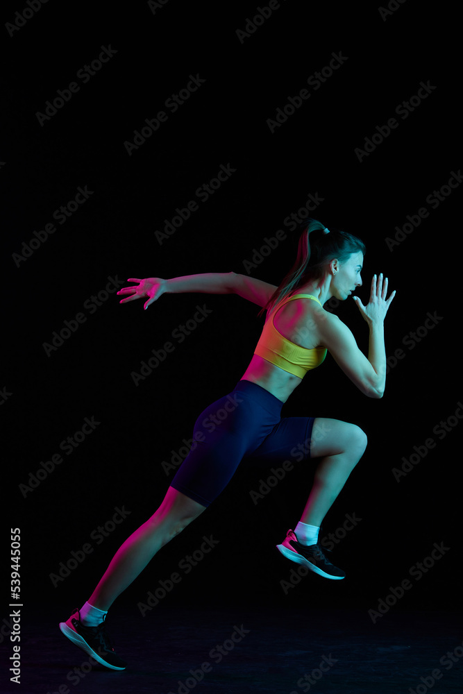 Fototapeta premium Dynamic portrait of young sportive woman training, running isolated on dark background in neon light. Competition, sport, fitness, speed and energy concept.