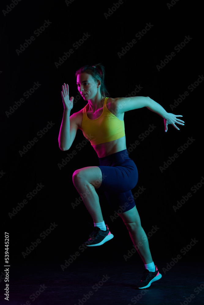Running technique. Professional female jogger running isolated on dark background in neon light. Sport, fitness, speed and energy concept.