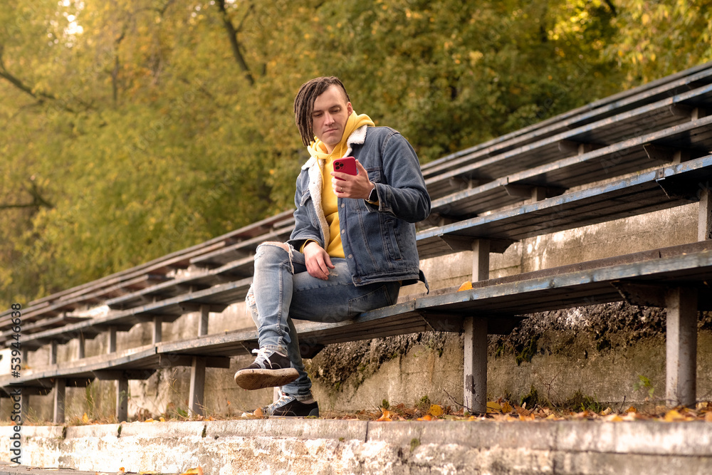 Fototapeta premium Young man with dreadlocks browsing mobile phone, sitting on old shabby bench of street bleacher.