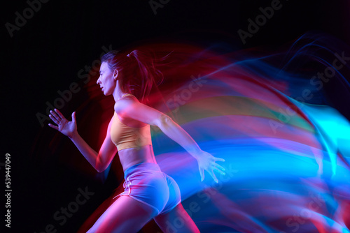 Energy. Professional female athlete, runner in motion over dark background in mixed neon light. Art, beauty, sport, cyberpunk concept