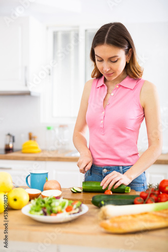 Positive young girl slicing vegetables for salad in cozy kitchen