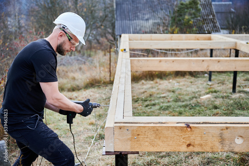 Man building frame house on pile foundation. Male worker drilling hole by electric drill in wooden frame of future house on construction site. Builder wearing helmet and goggles. Carpentry concept.