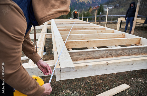 Man worker building wooden frame house on pile foundation. Carpenter using tape measure for measuring wooden planks. Carpentry concept.