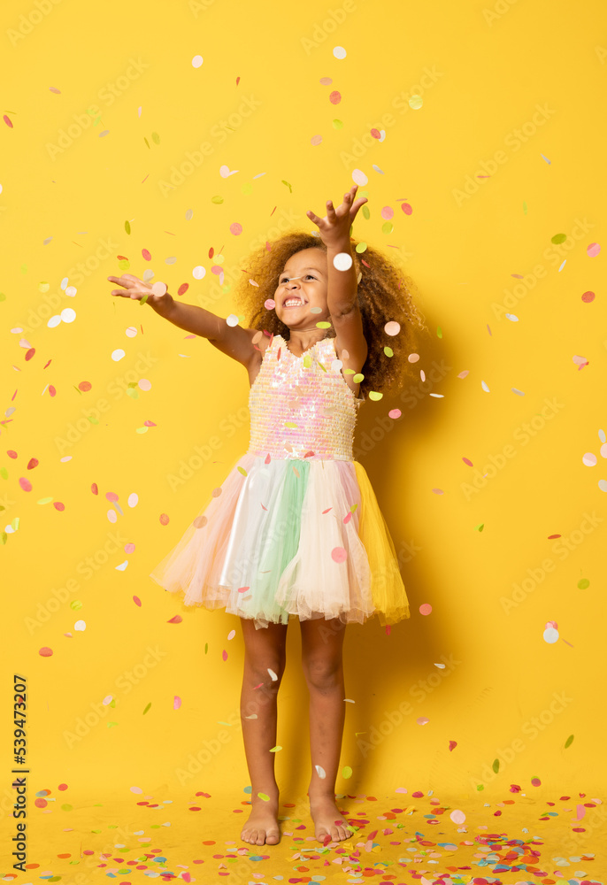 Funny little girl dressed for party in sequins and tutu blowing confetti . Isolated on yellow background. Funny little girl dressed for party in sequins and tutu blowing confetti . Isolated on yellow background.