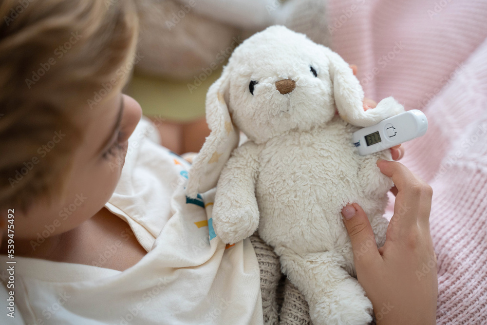 Sick child playing with his ill toy hare, measure body temperature with electronic thermometer. Role playing, child playing doctor with plush toy. Children cold and flu,  illness concept. Top view.