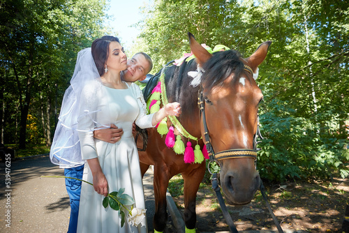 Stylish groom in a blue suit walk with the bride in a white dress in nature in the park with white horse. Wedding portrait of happy newlyweds on a background of greenery