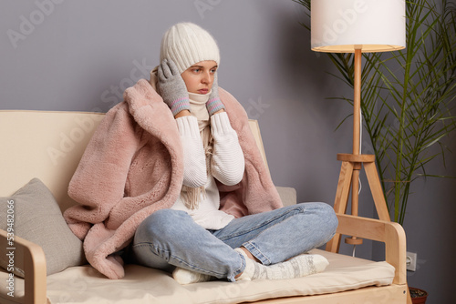 Indoor shot of sad unhappy woman wearing warm clothes and wrapped in scarf sitting on sofa in room, trying to get warm her ears, posing in house with turning off central heating.