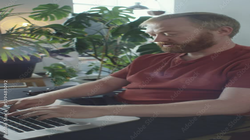 Vertical shot of redhead bearded man sitting on sofa at home and typing on laptop while working remotely Vertical shot of redhead bearded man sitting on sofa at home and typing on laptop while working remotely