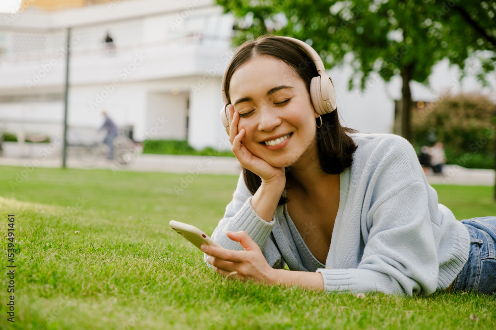 Obraz premium Young asian woman smiling and using cellphone while resting in park