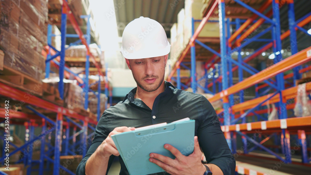 Close-up of Caucasian logistics employee examining the order of cargo cardboard boxes on paperwork. Young adult logistics manager is holding paperwork to record the stock of goods during work hours. Close-up of Caucasian logistics employee examining the order of cargo cardboard boxes on paperwork. Young adult logistics manager is holding paperwork to record the stock of goods during work hours.