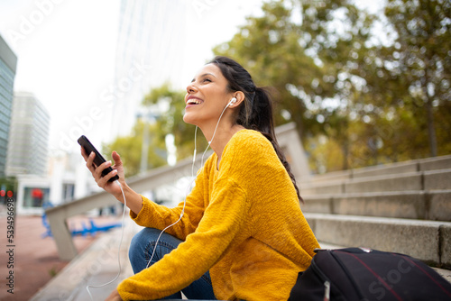 Side of young woman listening to music with earphones and cellphone in city photo