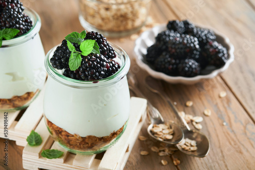 Yogurt with granola, blackberry berry fruits and muesli served in glass jar on wooden background. Healthy breakfast concept. Healthy food for breakfast, top view