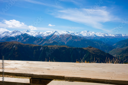 Close-up of an empty wooden bench against a blurred background. Edge of table on a sunny day in the mountains. Snow covered mountains and a blue sky. hike, break, view