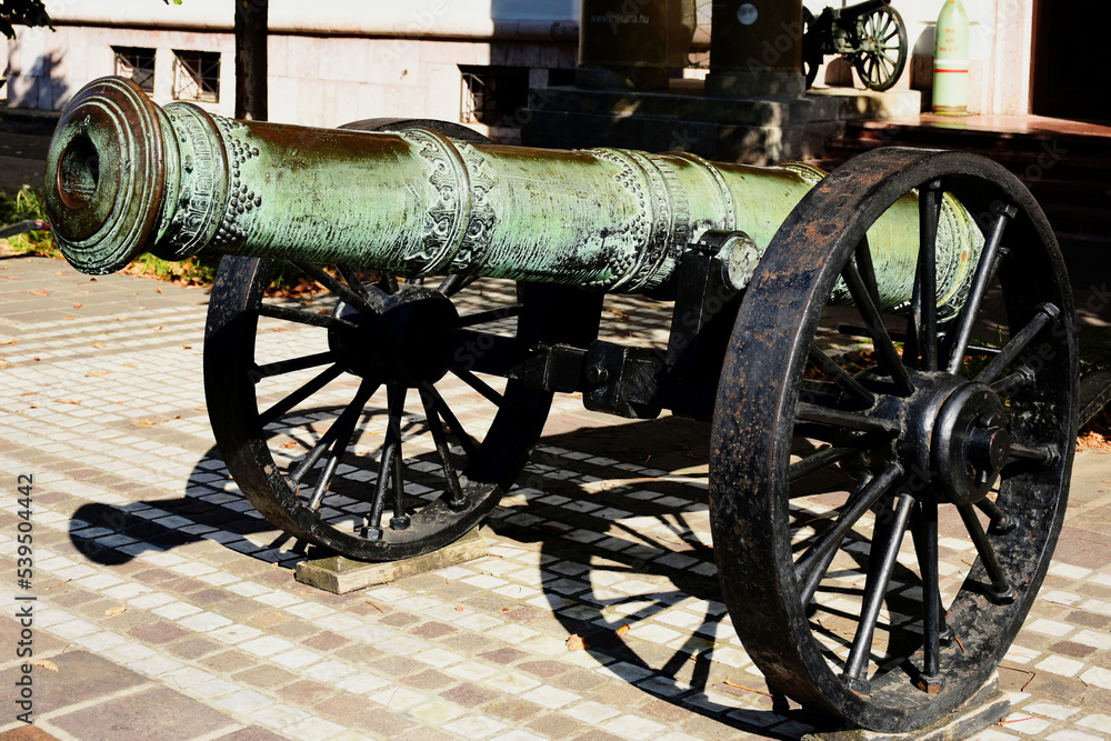 tarnished and aged old antique brass cannon closeup. diminishing perspective. large steel wheel. historic military equipment display. street setting. brick fortress wall. decorative pattern.