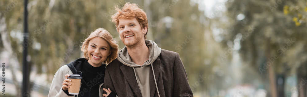 happy young woman in coat hugging man and walking with paper cup in autumnal park, banner. happy young woman in coat hugging man and walking with paper cup in autumnal park, banner.