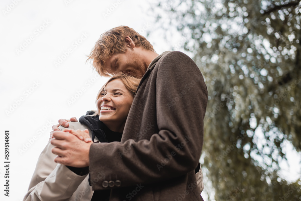 low angle view of redhead man kissing head of cheerful blonde woman during date in park. low angle view of redhead man kissing head of cheerful blonde woman during date in park.