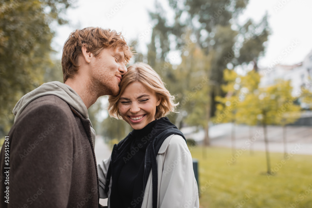 happy redhead man kissing head of blonde girlfriend smiling in park. happy redhead man kissing head of blonde girlfriend smiling in park.