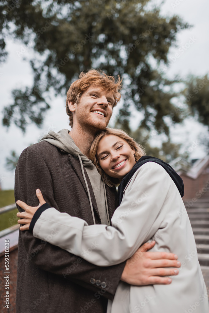 happy young woman hugging redhead boyfriend in autumnal coat in park. happy young woman hugging redhead boyfriend in autumnal coat in park.