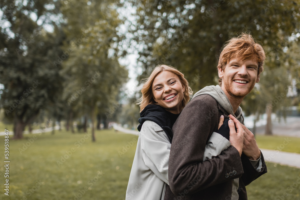 positive young woman in coat hugging cheerful redhead boyfriend in park.