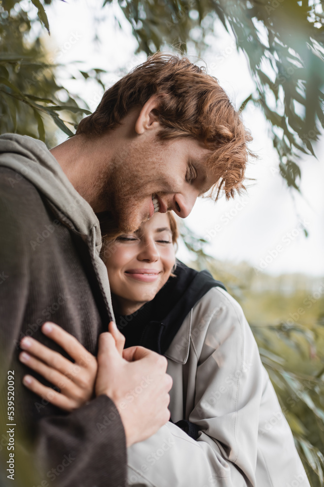 pleased blonde woman hugging joyful young man near green leaves on tree. pleased blonde woman hugging joyful young man near green leaves on tree.