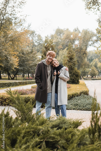 joyful and young man in coat hugging blonde girlfriend while standing around plants in park. © LIGHTFIELD STUDIOS