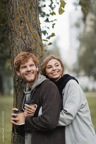 happy blonde woman holding paper cup and hugging with boyfriend near tree trunk.