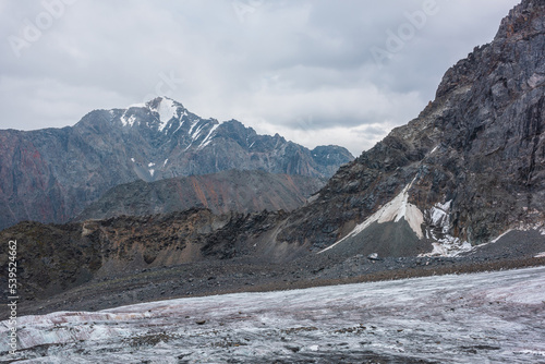 Awesome landscape with long glacier tongue against high mountain range with sharp rocky peaked top in overcast weather. Cone shaped pointy peak in dramatic cloudy sky. Large glacier among sharp rocks.