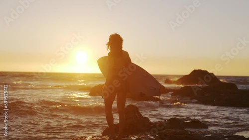 Young female surfer standing on a rock under the sunset sky holding a surfboard watching the ocean waves rise. 