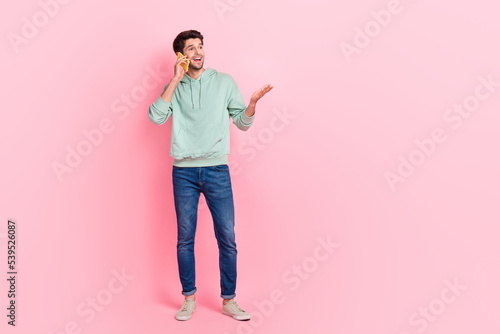 Full length photo of positive man wear stylish hoodie look empty space speak modern device gadget space isolated on pink color background