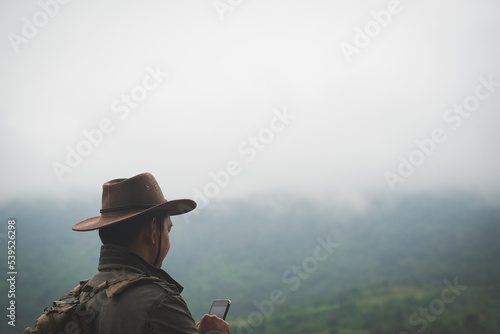 Freedom traveler man in hat carrying a backpack stands at the top of a mountain and using a smartphone on a foggy day.Adventure travel and success concept