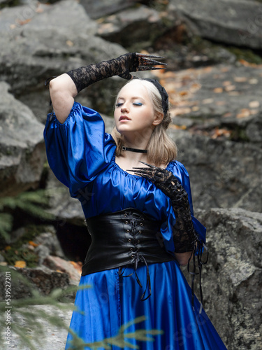 A beautiful woman in a long blue dress, black leather corset and black lace gloves surrounded by rocks. Forest Nymph.