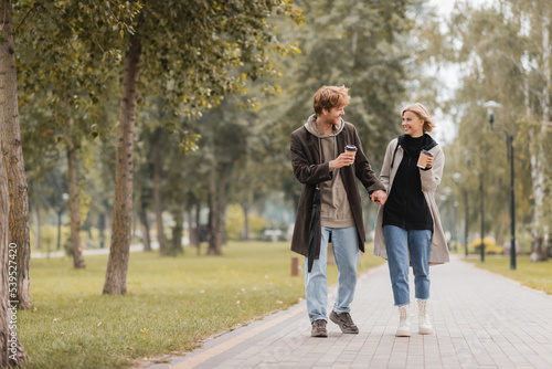 full length of happy couple in coats holding hands and walking with coffee to go in autumnal park. © LIGHTFIELD STUDIOS