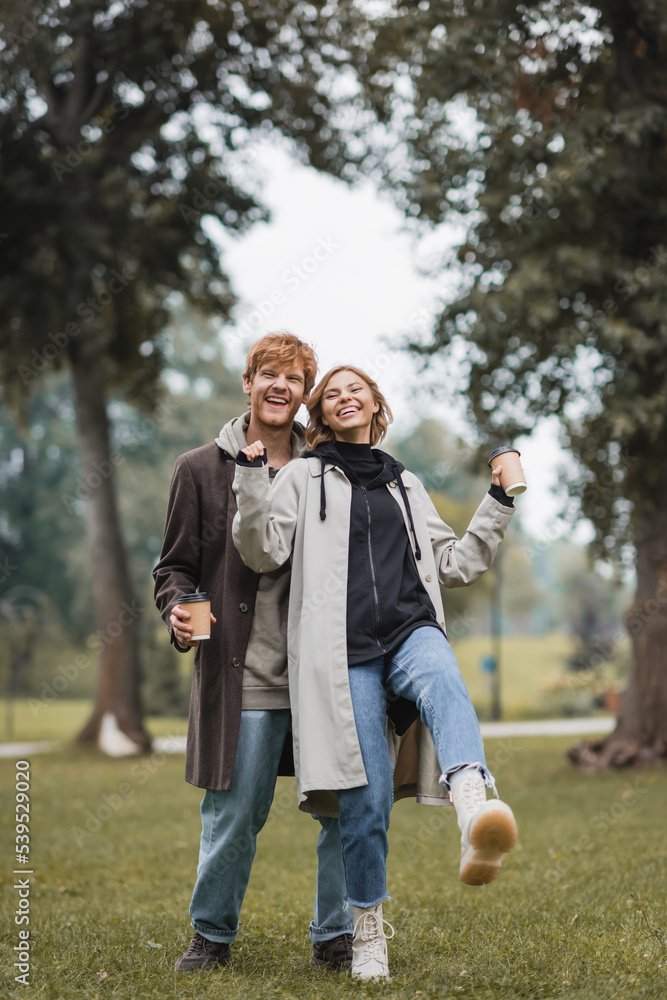 Fototapeta premium full length of happy redhead man and pleased young woman holding coffee to go while walking in park.