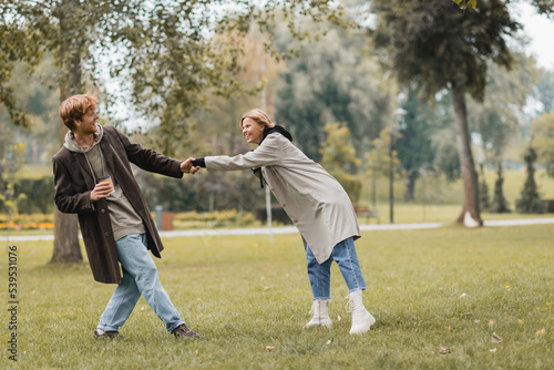 full length of happy young man in coat holding paper cup and pulling girlfriend in autumnal park.