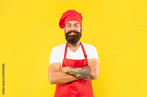 Happy man cook in red apron and toque keeping arms crossed yellow background, confidence