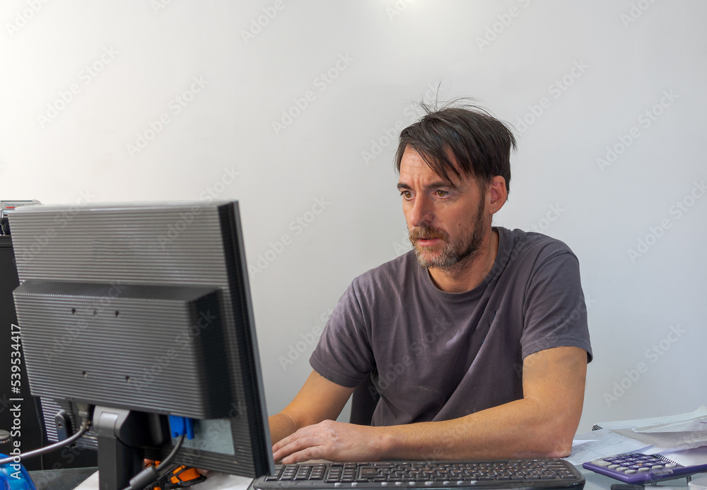 The chief mechanic and owner of his business, sitting in his office, in front of his computer screen and keyboard, reviews customer invoices and repairs for the current month in his workshop in Granad The chief mechanic and owner of his business, sitting in his office, in front of his computer screen and keyboard, reviews customer invoices and repairs for the current month in his workshop in Granad