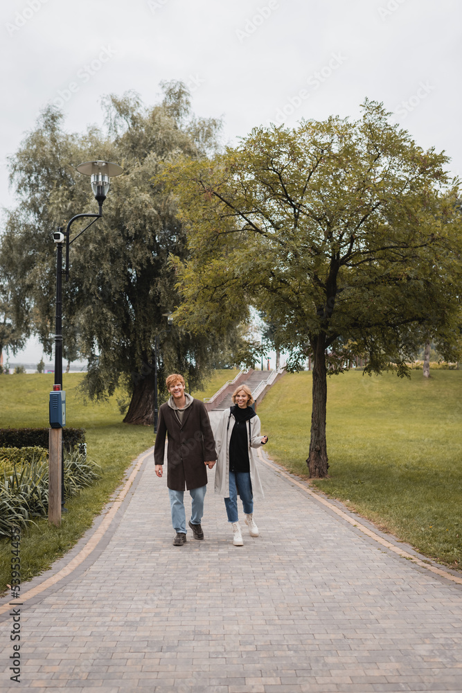 full length of joyful redhead man and pleased young woman in coat holding hands while walking in park. full length of joyful redhead man and pleased young woman in coat holding hands while walking in park.