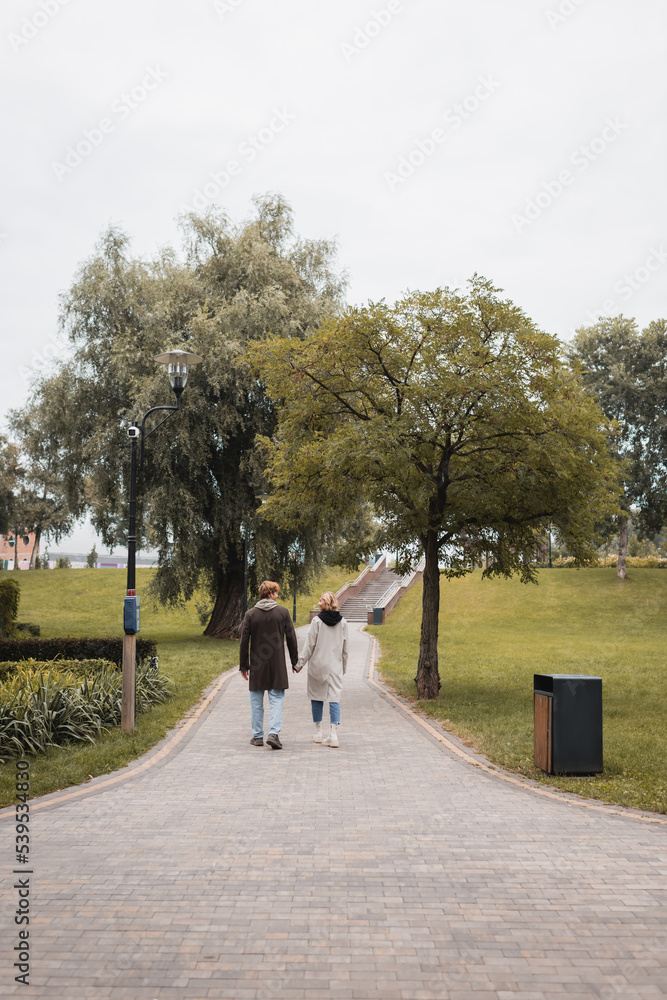 back view of happy redhead man and pleased young woman holding hands while walking in park. back view of happy redhead man and pleased young woman holding hands while walking in park.