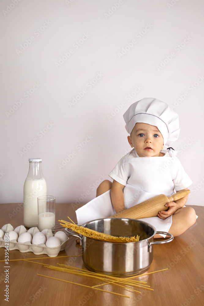A cook boy in an apron and a cap sits on a table with a pot of spaghetti, a rolling pin, milk and eggs. The concept of a holiday, food, course, training, advertising, announcement, promotion, postcard