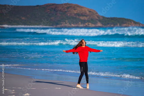 A beautiful long-haired girl in a red sweatshirt walks on the sand at the famous lucky bay beach at sunset; a happy girl on the beach with white sand, turquoise water and hills in the background