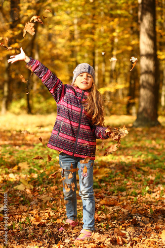 Sunlit portrait of a cheerful girl throwing leaves in the autumn park