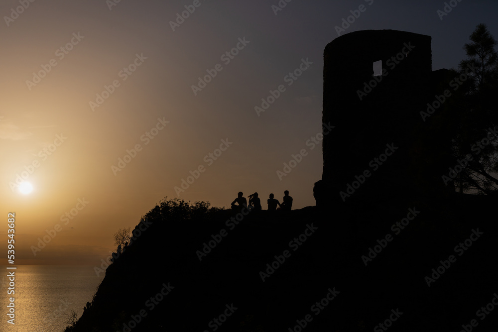 Grupo de amigos jóvenes contemplando la puesta de sol y divirtiéndose junto a la Torre des Verger, un mirador junto al mar en las montañas de la Serra de Tramuntana de Mallorca.
