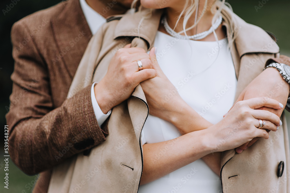 Fototapeta premium A stylish man in a brown suit gently hugs a girl in a white dress, beige coat outdoors in autumn, holding hands. Wedding photography of the newlyweds.