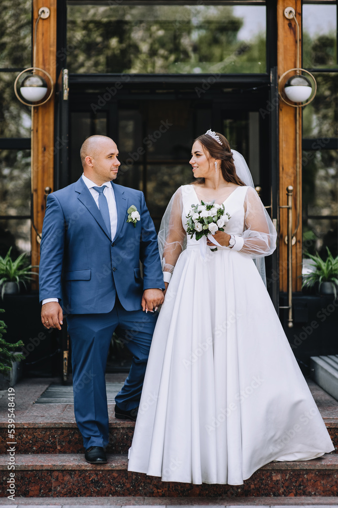Fototapeta premium An adult stylish groom in a blue suit and a beautiful smiling bride in a white dress hold hands, against the backdrop of a city street in a park. Wedding photography of smiling newlyweds, portrait.