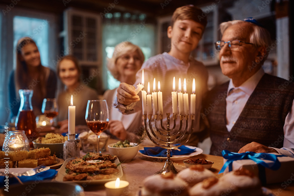Close up of Jewish boy lighting candles in menorah while celebrating Hanukkah with his family at dining table. Close up of Jewish boy lighting candles in menorah while celebrating Hanukkah with his family at dining table.